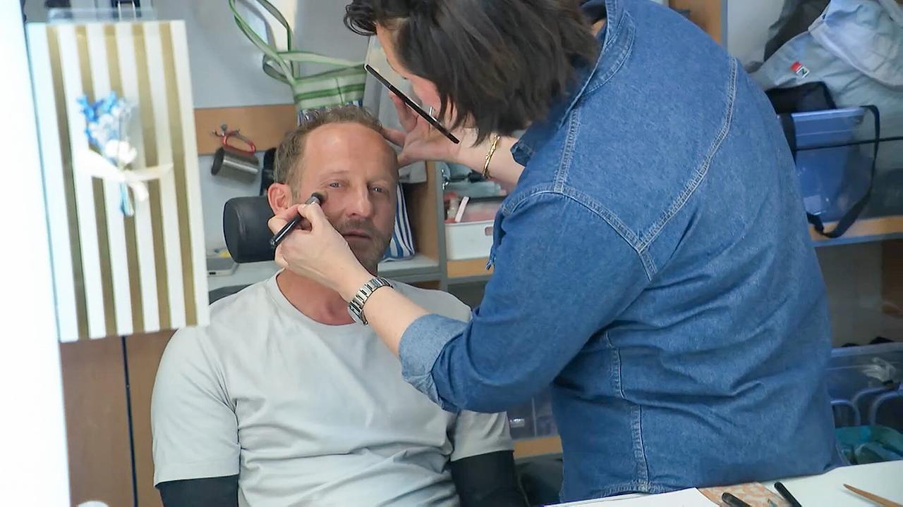 Maximilian Brückner in der Maske wird von einer Frau geschminkt.