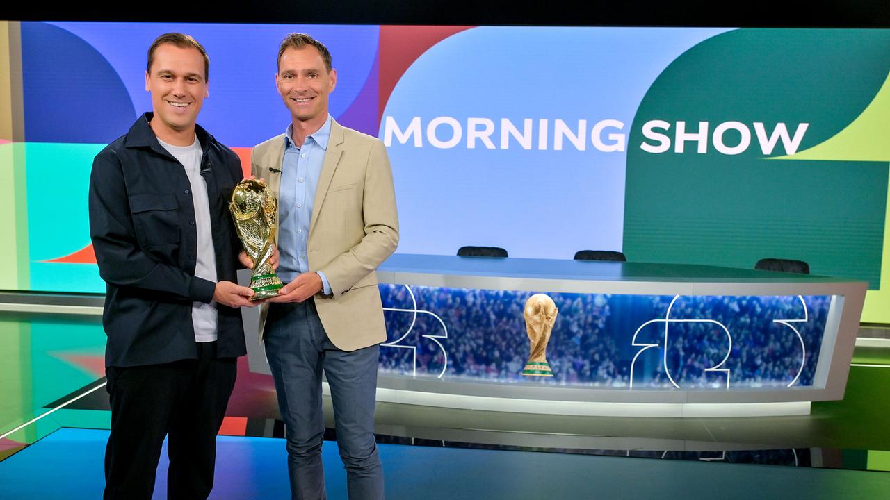 © ORF/Thomas Ramstorfer Michael Liendl und Christian Diendorfer stehen in einem modernen „Morning Show“-TV‑Studio und halten gemeinsam den goldenen Fußball‑WM-Pokal, während im Hintergrund der Studiotisch und die bunte Kulisse zu sehen sind.