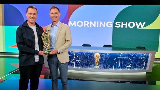 © ORF/Thomas Ramstorfer Michael Liendl und Christian Diendorfer stehen in einem modernen „Morning Show“-TV‑Studio und halten gemeinsam den goldenen Fußball‑WM-Pokal, während im Hintergrund der Studiotisch und die bunte Kulisse zu sehen sind.