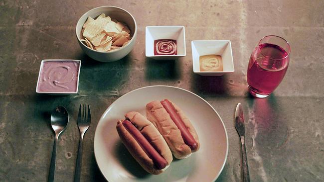 Ein gedeckter Platz am Tisch - zwei Hot-Dog-Brötchen auf dem Teller, Kartoffel-Chips, lilafarbene Sauce, Ketchup und Senf in Schalen, dazu ein Glas roten Safts