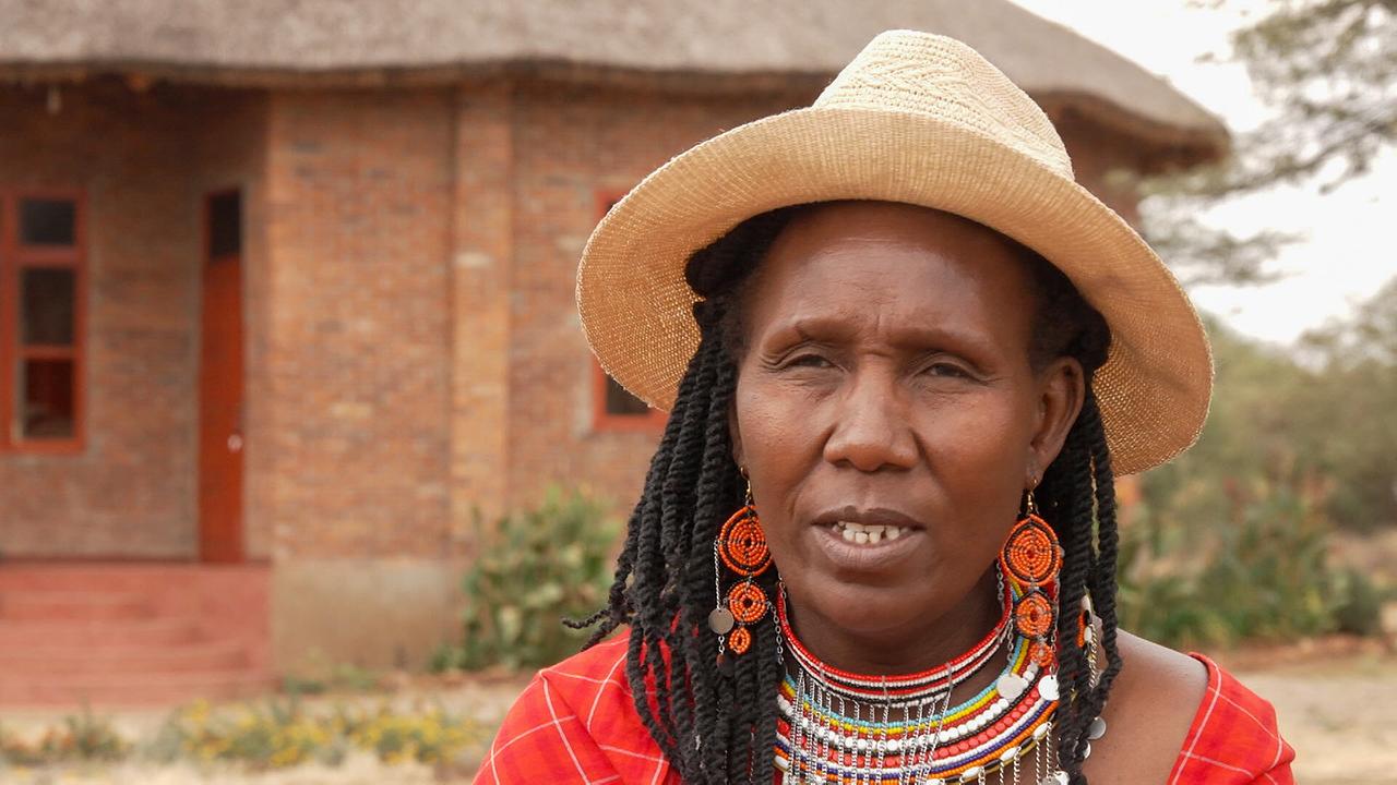 Profil von Massai Maanda Ngoitiko mit Hut und traditionellem Schmuck vor einem Backsteingebäude