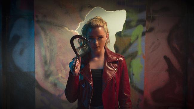 Kathryn Newton als Millie Kessler mit schwarzem Top und roter Lederjacke steht se vor einer bunt bemalten Wand, die durchbrochen worden ist, sie hält eine Sichel in der Hand und blickt entschlossen drein