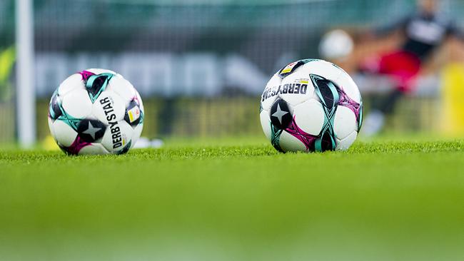 Drei Fußbälle liegen auf dem Rasen vor einem Tor, im Hintergrund ist unscharf ein Spieler in Aktion zu sehen.