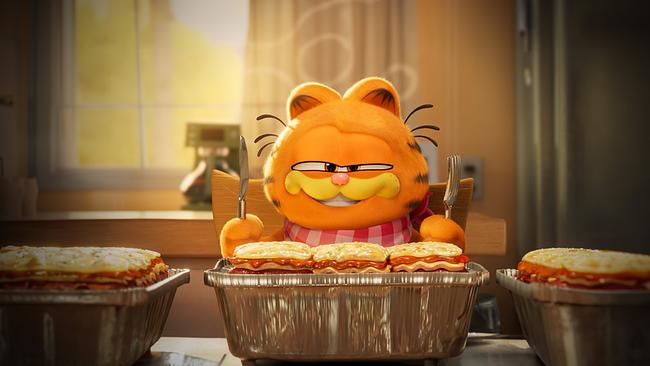 Der orangefarbene, getigerte Kater Garfield sitzt mit Messer und Gabel lächelnd am Tisch, auf dem köstliche Lasagne zum Verzehr bereitstehen