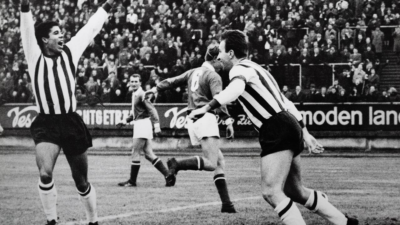 Eine historische Schwarz‑Weiß‑Aufnahme zeigt eine Fußballszene im Stadion, in der Spieler u.a. Helmut Köglberger jubelnd mit erhobenen Armen vor dicht gedrängten Zuschauerrängen agieren.