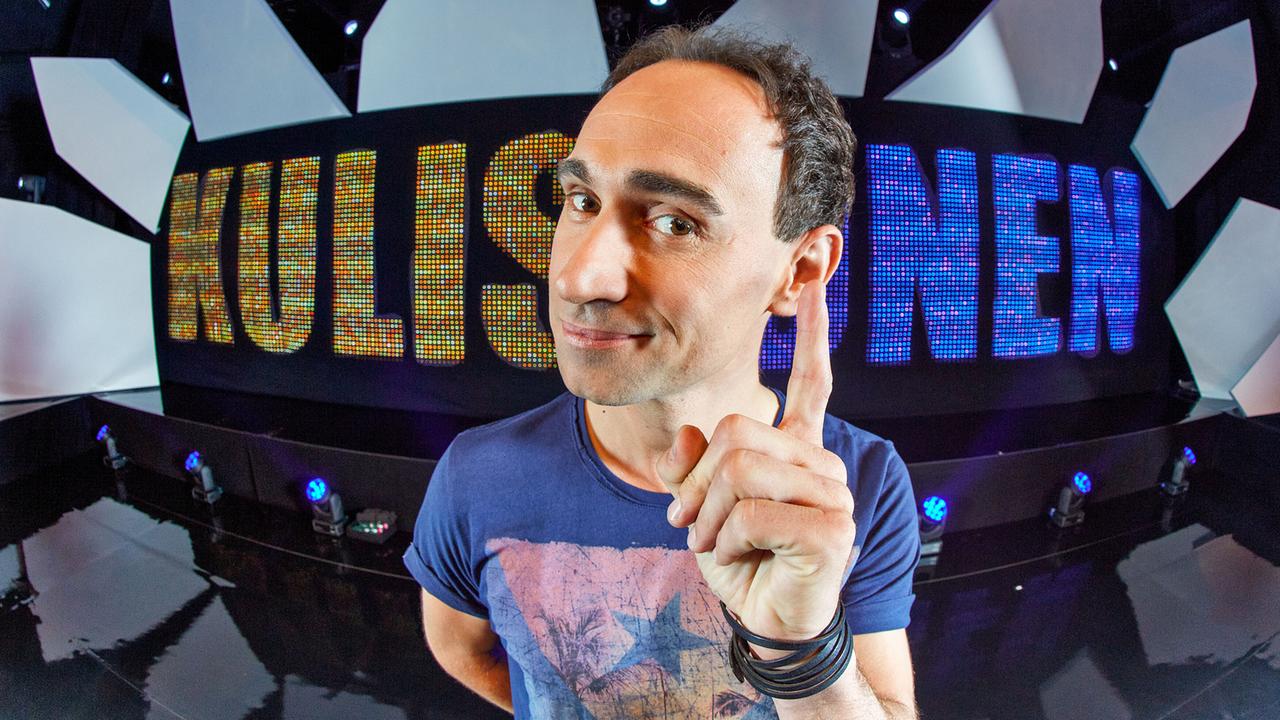 © ORF/Hoanzl/Markus Wache Gernot Kulis steht auf einer "Kulisionen"-Bühne und blickt mit erhobenem Zeigefinger in die Kamera.