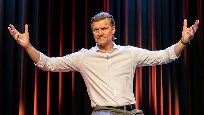 Gery Seidl in weißem Hemd gestikuliert mit weit ausgebreiteten Armen auf der Bühne.