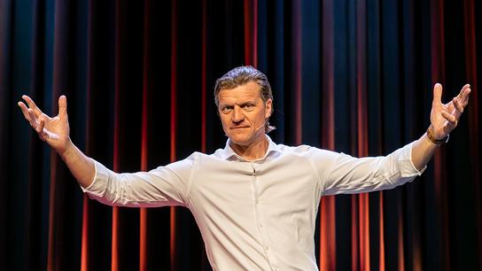 Gery Seidl in weißem Hemd gestikuliert mit weit ausgebreiteten Armen auf der Bühne.