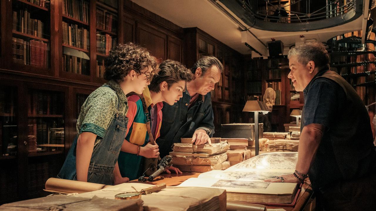 © ORF/Sony Pictures/CTMG/Jaap Buitendijk Zwei Jugendliche und ein Mann betrachten in einem Antiquariat ein großes Buch, ein weiterer Mann ihnen gegenüber erklärt etwas zur aufgeschlagenen Seite