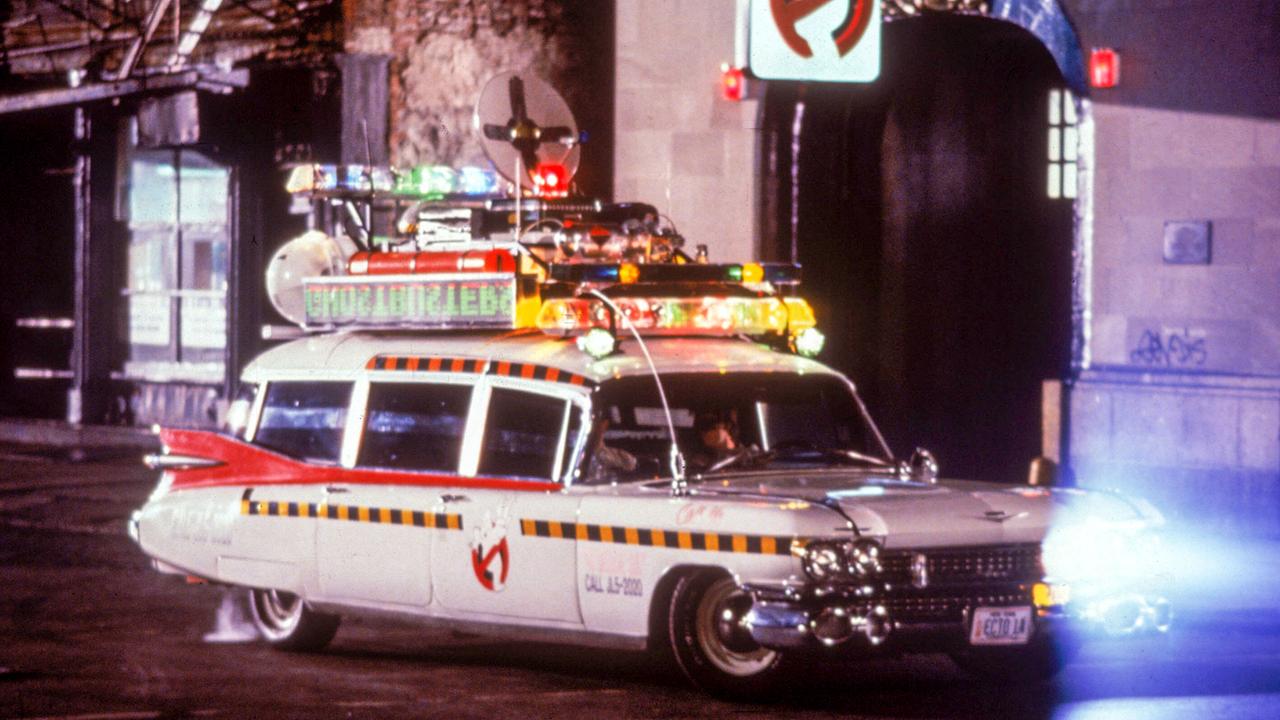 Das Auto der Ghostbusters im Einsatz in einer Straße in der Nacht, ein weißer Kombi mit bunt leuchtenden technischen Apparaten am Dach