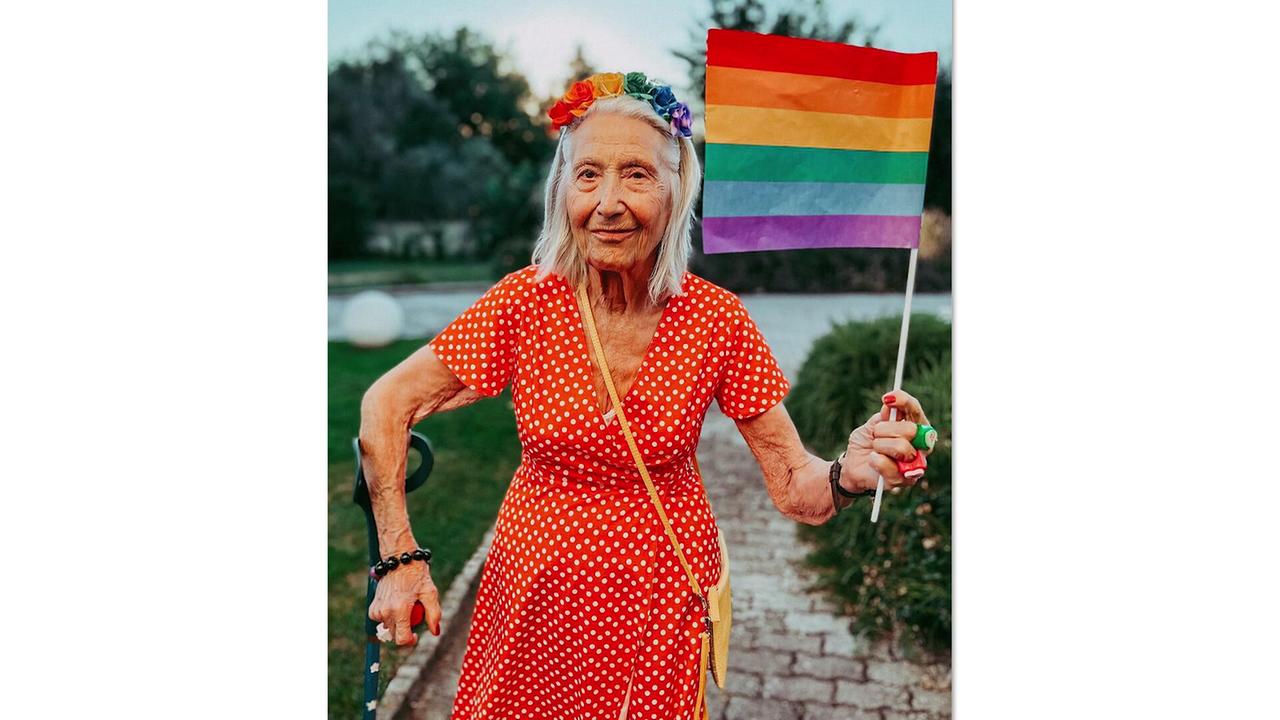 Italiens bekannteste Insta-Großmutter: Licia Fertz, in einem roten, gemusterten Kleid und regenbogenfarbigen Haarreifen hält sie die Regenbogenfahne in der Hand