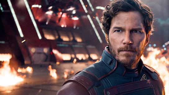 Chris Pratt als Star-Lord, mit Bart und gewellten mittellangen Haaren steht er in dunklem blau-rotem, ledernen Anzug in einem Raum, i dem es hinter ihm brennt, aufmerksam blickt er nach vorne