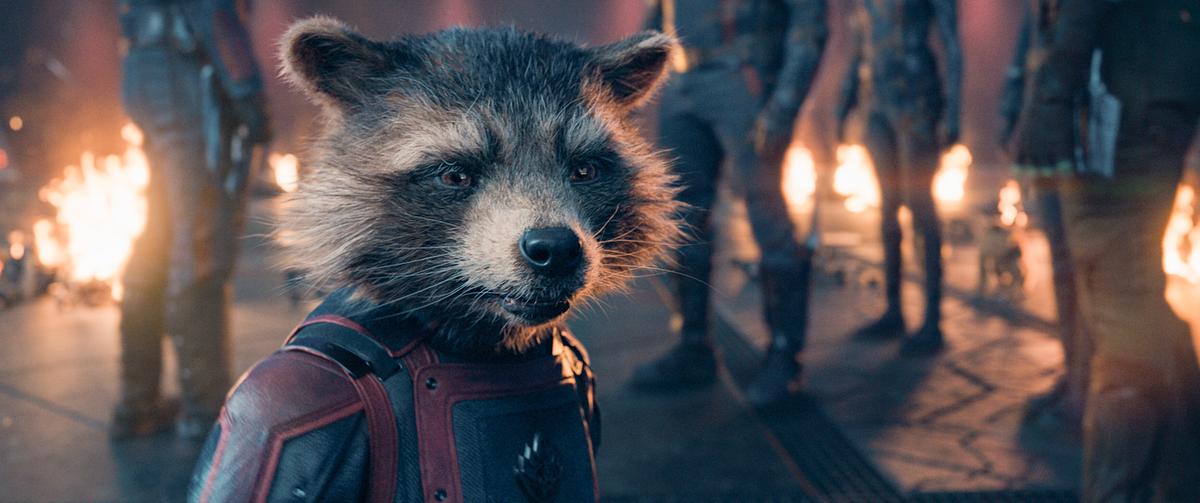© ORF/Disney/© 2023 MARVEL Der Waschbär Rocket steht in seinem blau-rotem Anzug in einem Transportraum, im Hintergrund sind weitere Personen zu sehen, dahinter brennt ein Feuer
