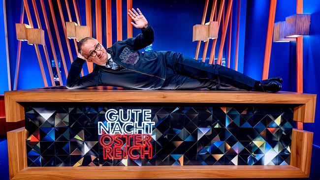 © ORF/Hans Leitner Peter Klien liegt seitlich auf seinem Moderationstisch im "Gute Nacht Österreich"-Studio und winkt in die Kamera.