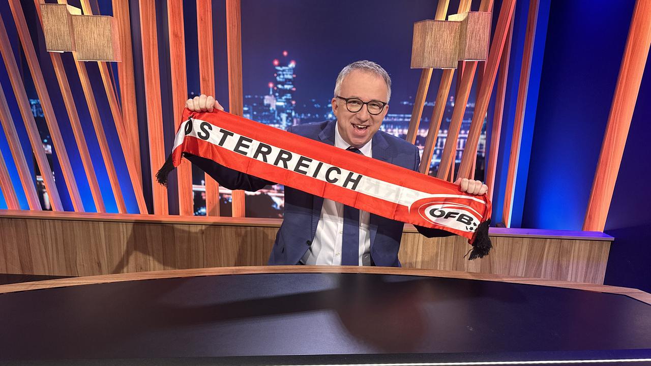 Peter Klien im "Gute Nacht Österreich"-Studio hält einen rot-weißen ÖFB-Schal mit der Aufschrift „Österreich“ hoch.