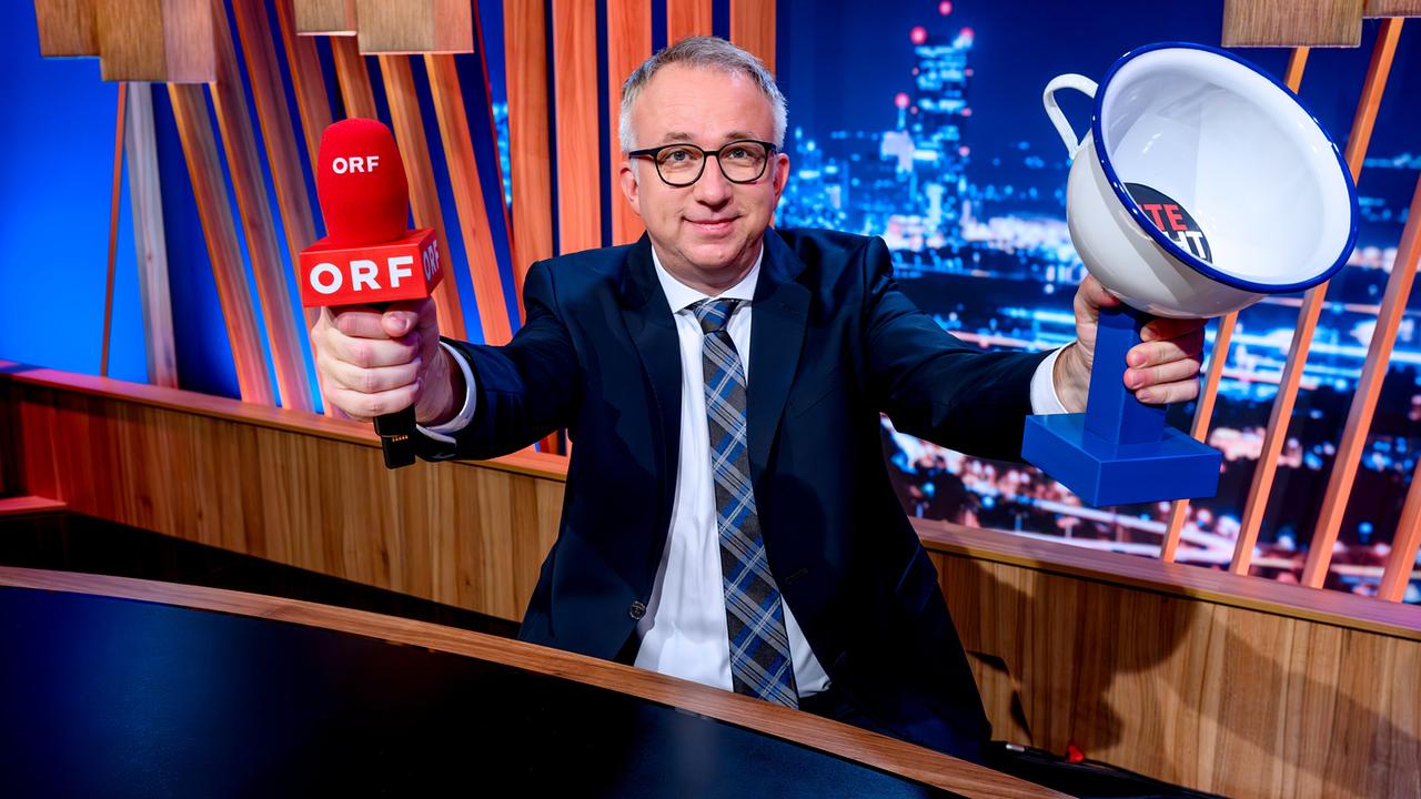 Peter Klien sitzt im Studio und hält seine Arme hoch, während er in der rechten Hand ein ORF-Mikro und in der linken Hand den "Gute Nacht Österreich - Der Gute Nacht-Topf" hält.