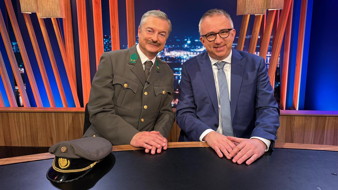 Alfred Dorfer in Militäruniform und Peter Klien im Sakko sitzen am Moderatorenpult im "Gute Nacht Österreich"-Studio.