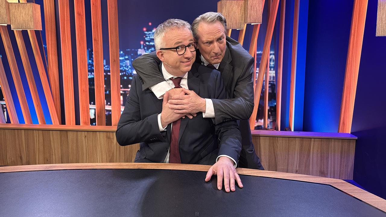 Robert Palfrader umarmt Peter Klien, der am Moderatorenpult sitzt, scherzhaft von hinten im "Gute Nacht Österreich"-Studio