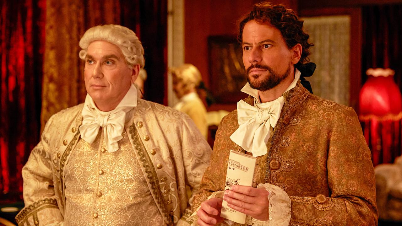 Bryan Nichols und Harrow in historischer Kleidung, Nichols trägt eine weiße Perrücke, Harrow hält einen Folder in der Hand, beide schauen fokussiert ihr Gegenüber an