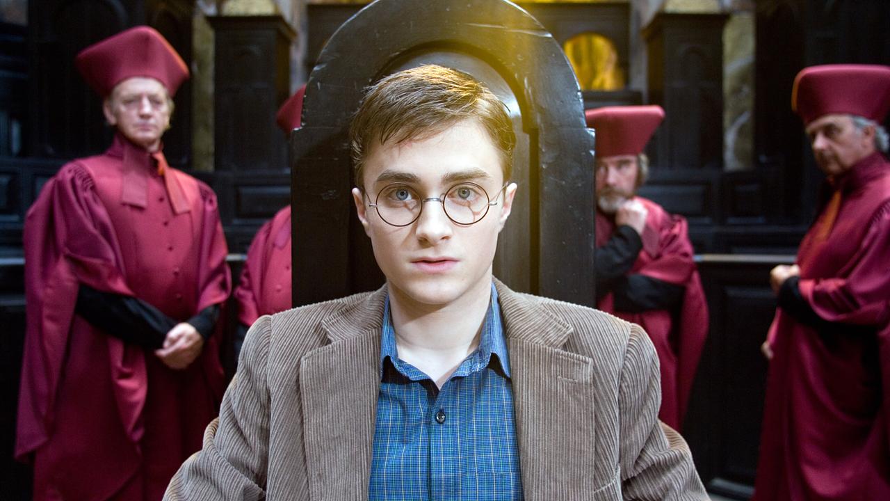 Harry Potter sitzt angespannt auf einem Stuhl mit hoher Lehne, hinter ihm sind drei Männer in purpurner Tracht eines geistlichen Ordens