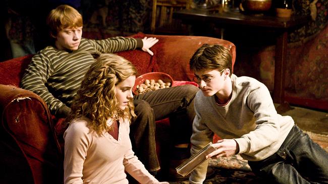 Ron sitzt in einem alten roten Sofa, vor ihm auf dem Boden sitzen Hermine und Harry Potter, alle schauen auf ein altes Buch, das Harry in der Hand hält
