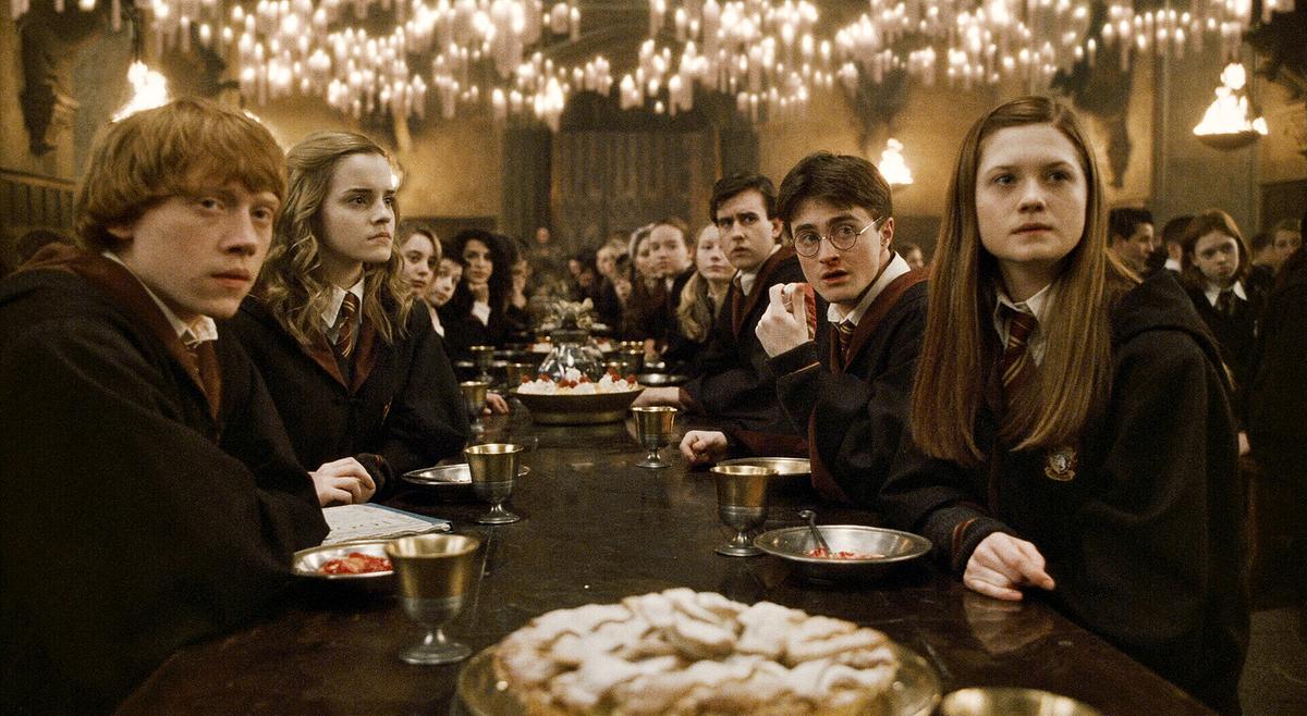© ORF/Warner Bros. Pictures Ron, Hermine, Harry udn Ginny sitzen mit ihren Mitschülerinnen und Mitschülern in Schuluniform beim Essen im Speisesaal von Hogwarts - alle blicken aufmerksam in dieselbe Richtung