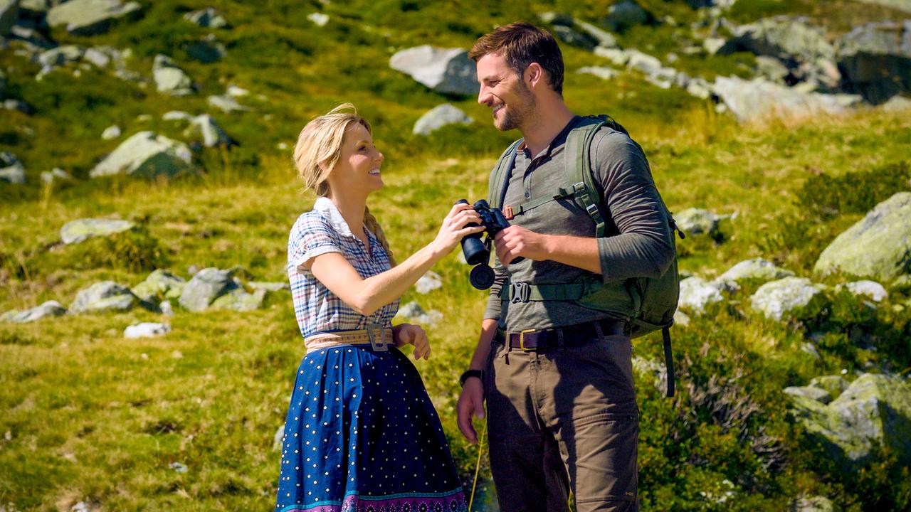 Melanie und Ben, der ihr ein Fernglas reicht, stehen lächelnd auf einer Almwiese.