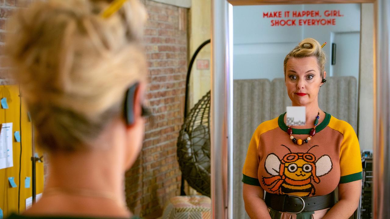 Nina, in einem gelb-orange-grünen Pullover mit einem Bienenmotiv steht in einem hellen Raum vor einem Spiegel mit der Aufschrift "Make it happen, Girl, shock everyone".