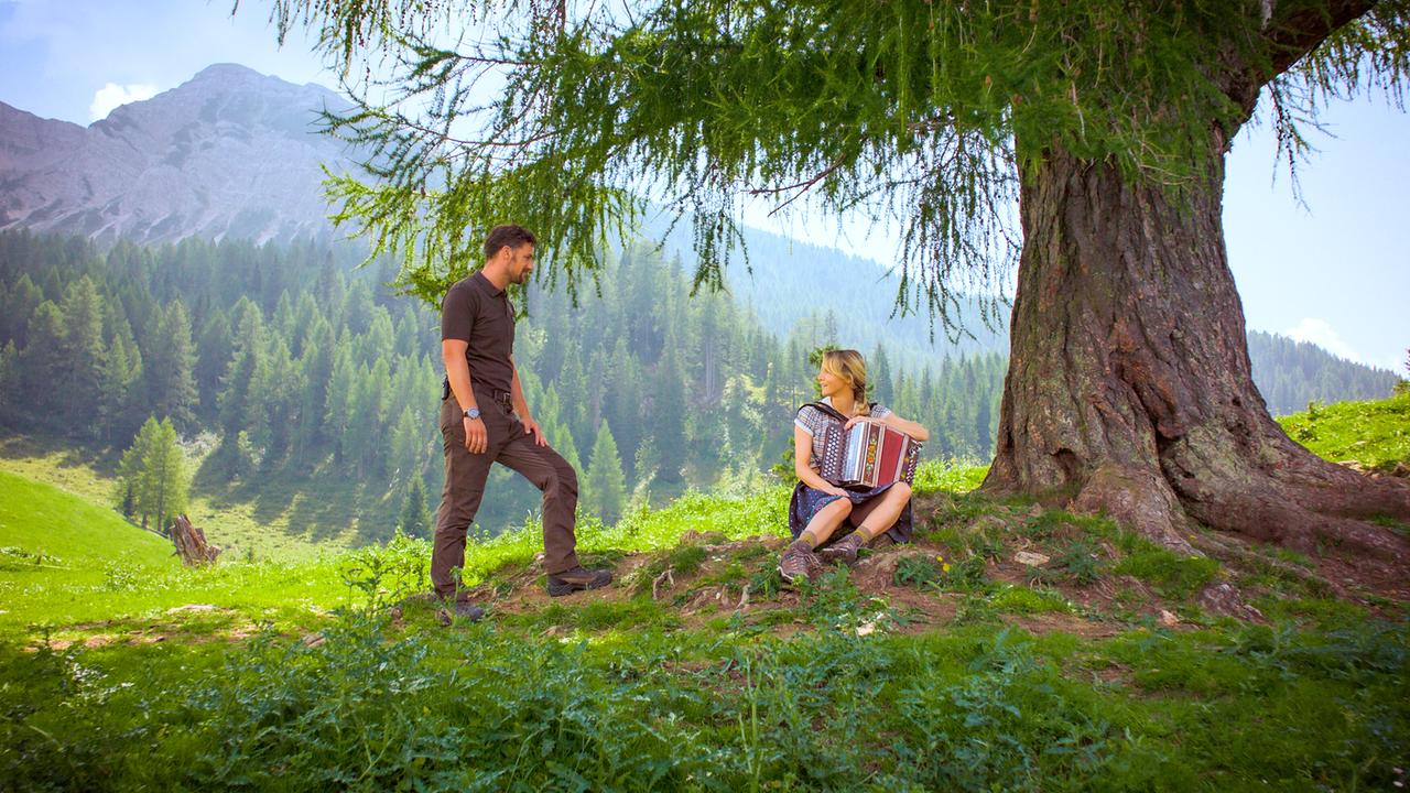 Unter einem großen Baum in den Bergen sitzt Melanie mit Ziehharmonika, während Ben neben ihr steht und sie anschaut.