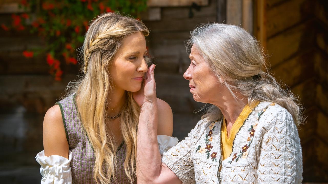 Melanie und Annabell sitzen in Trachtenkleidung vor einem Holzhaus und blicken sich vertraut an.