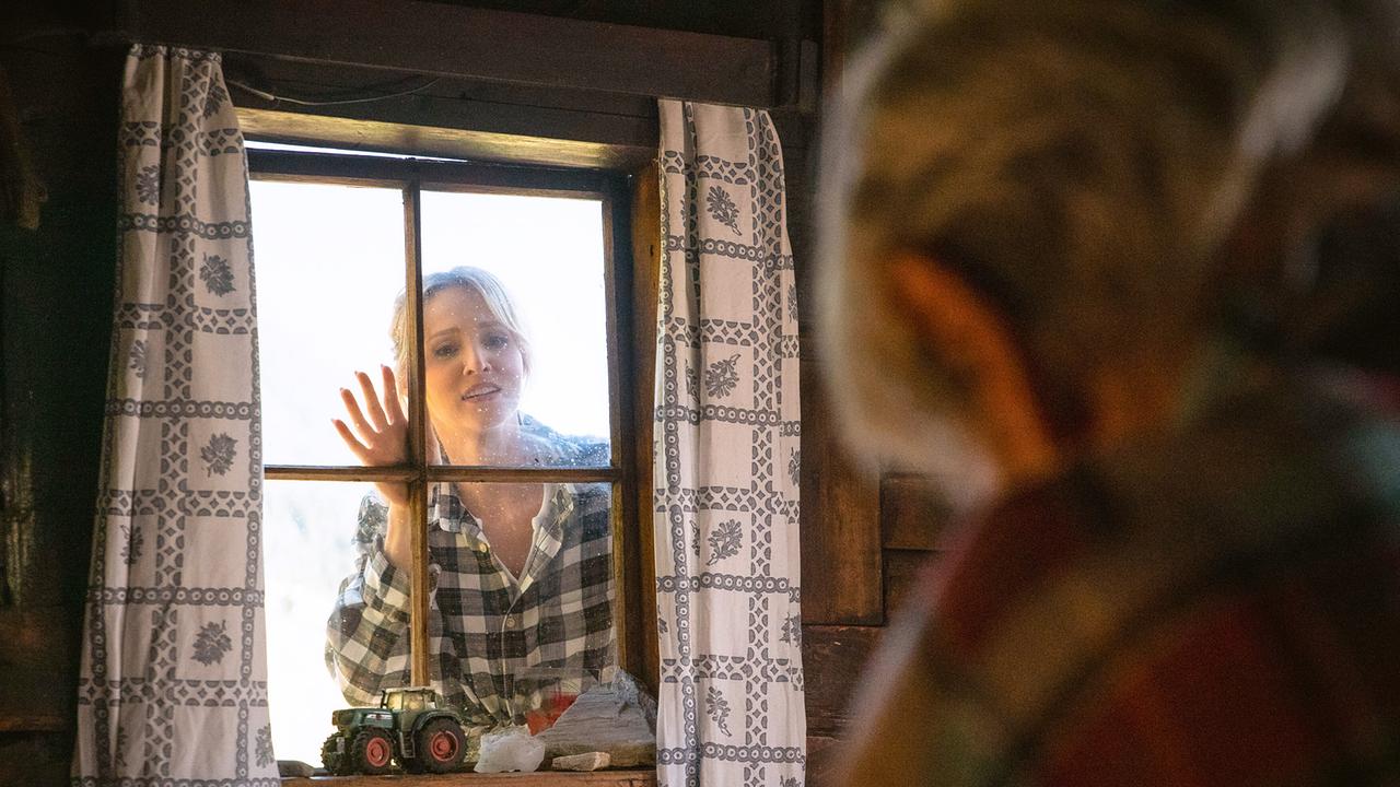 Melanie blickt von außen durch ein Fenster auf Richard in einer rustikalen Stube mit Vorhängen und einem Spielzeugtraktor auf der Fensterbank.