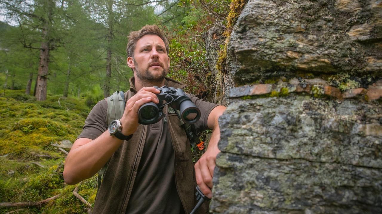 Ben lehnt mit einem Fernglas in der Hand an einem Felsen im Wald.