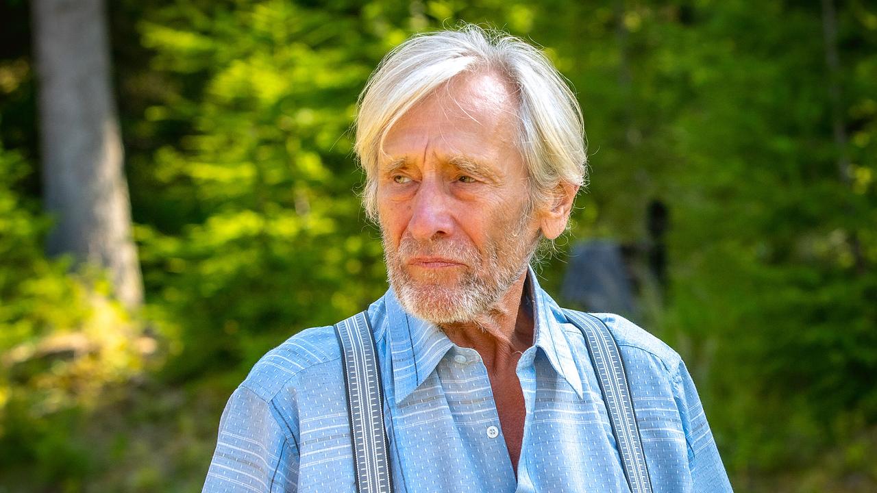 Der ältere Richard steht bei Sonnenschein auf einem Waldweg und schaut nachdenklich nach vorne.