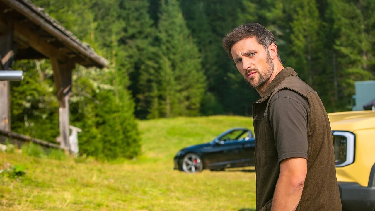Ben steht auf einer Almwiese nahe einer Hütte vor seinem gelben Pick-up und dreht sich um, während im Hintergrund ein Cabrio bei der Hütte geparkt ist.