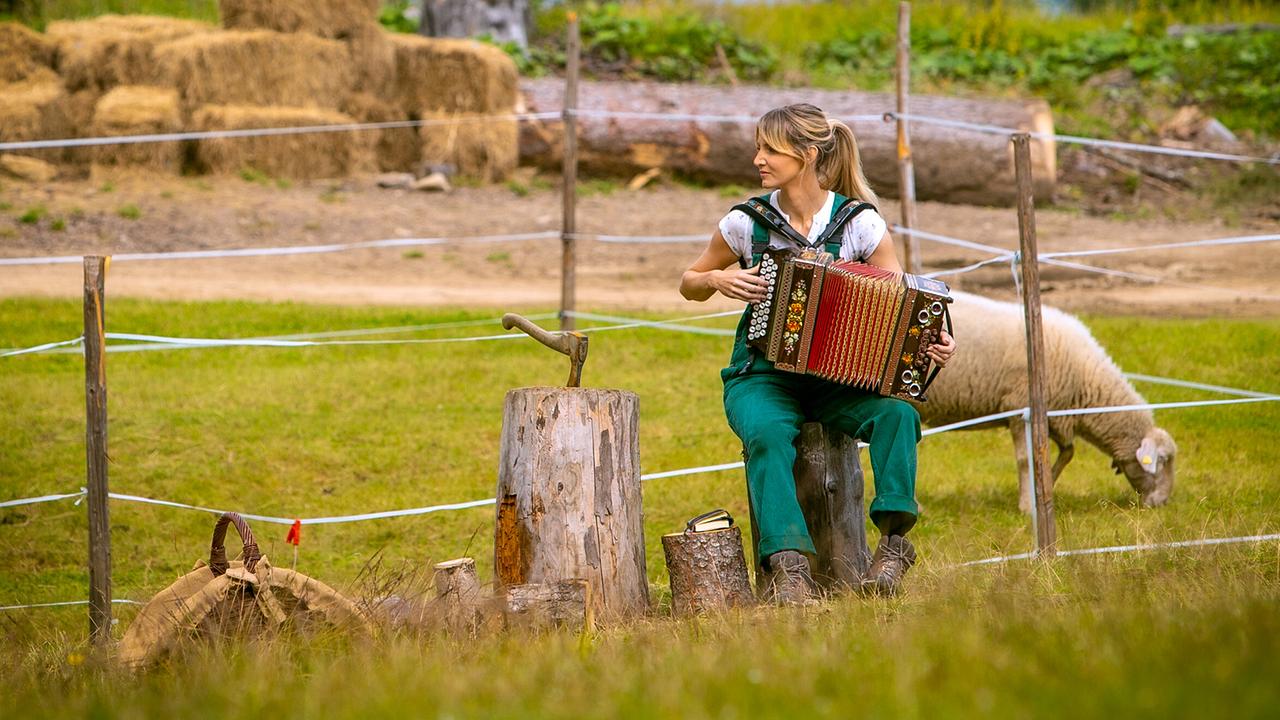 Melanie in einer grünen Latzhose sitzt auf einem Holzstumpf und spielt auf einer Wiese vor einem eingezäunten Bereich Ziehharmonika. Im Hintergrund ist ein Schaf zu sehen.