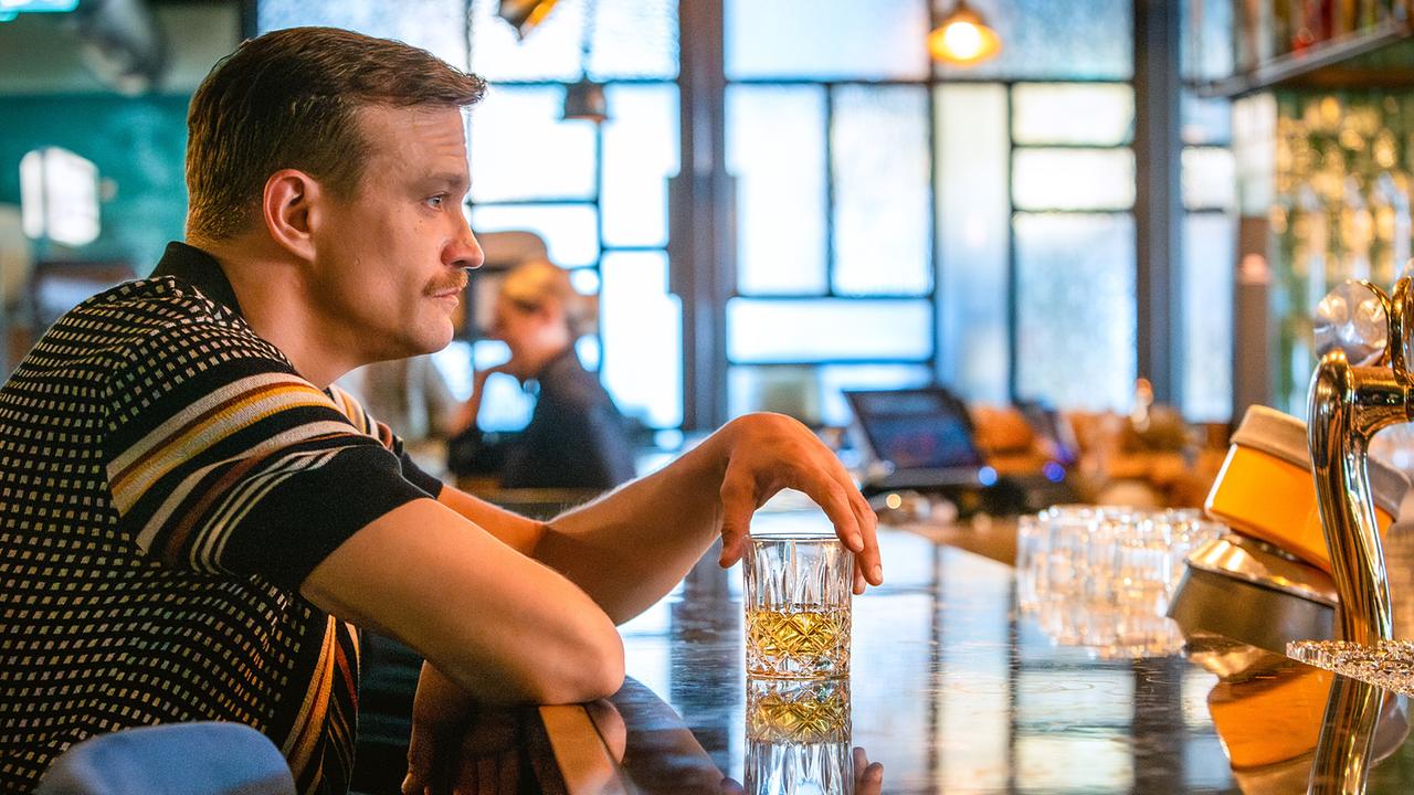 Marc sitzt an einer Bar und hält nachdenklich ein Glas mit einem Getränk in der Hand.