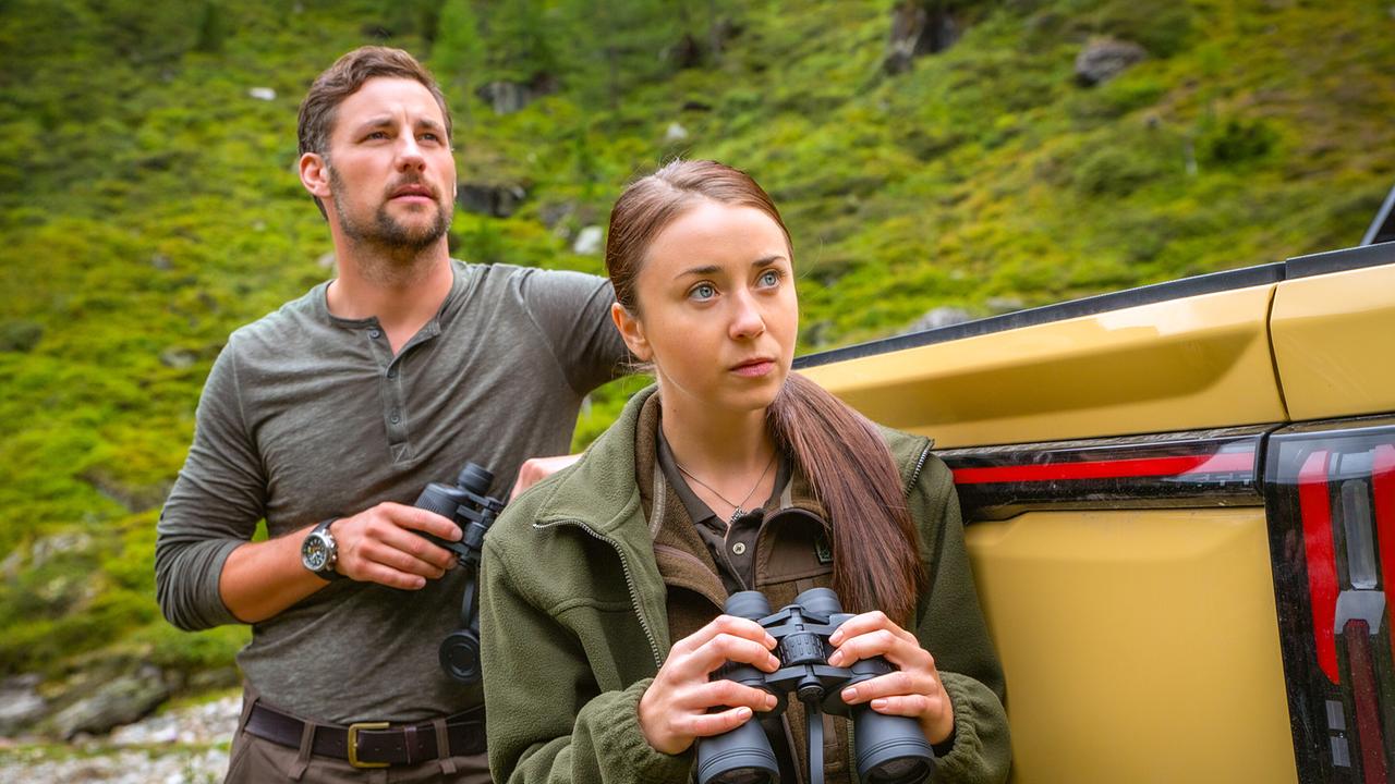 Ben und Heidi stehen mit Ferngläsern neben einem gelben Pick-up in einer bergigen Landschaft.