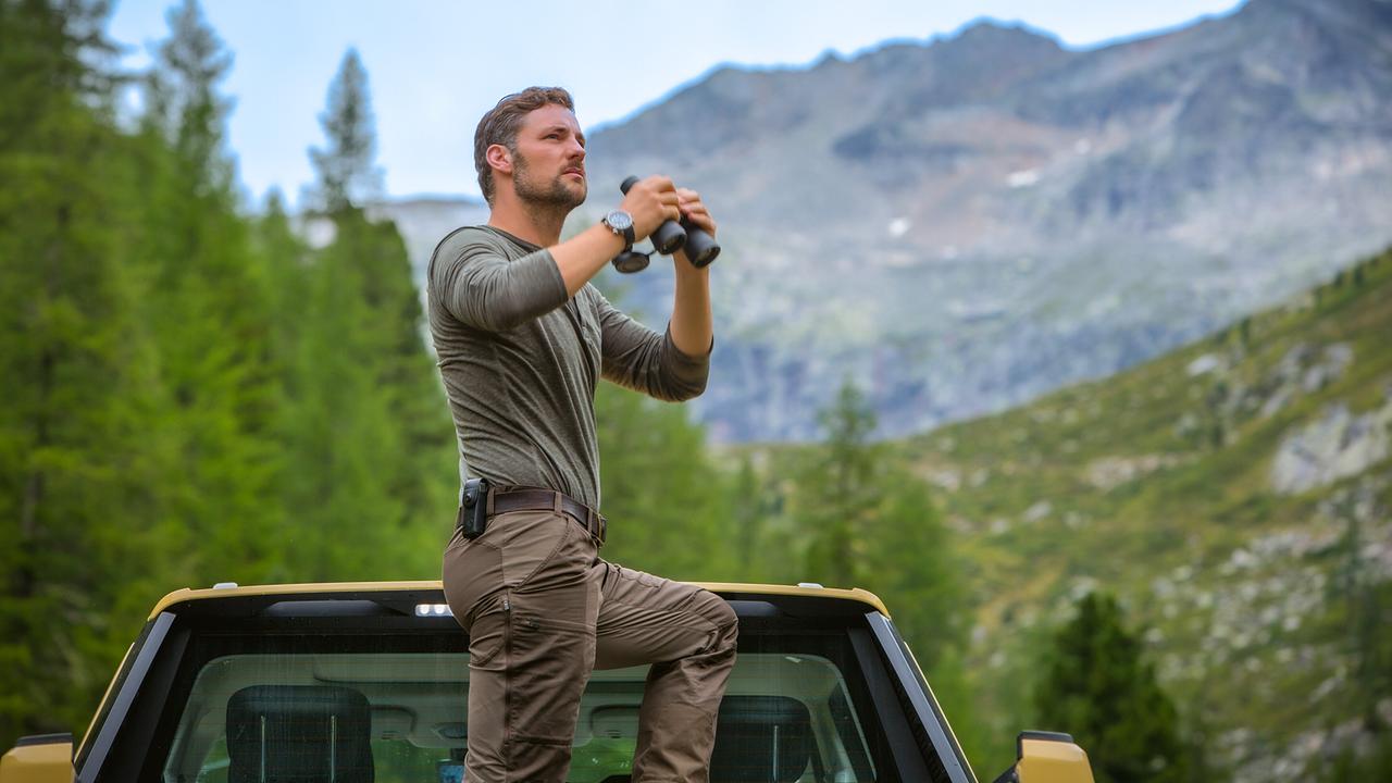 Ben steht auf der Ladefläche eines gelben Pick-ups mit Fernglas in der Hand in einer bergigen Landschaft.