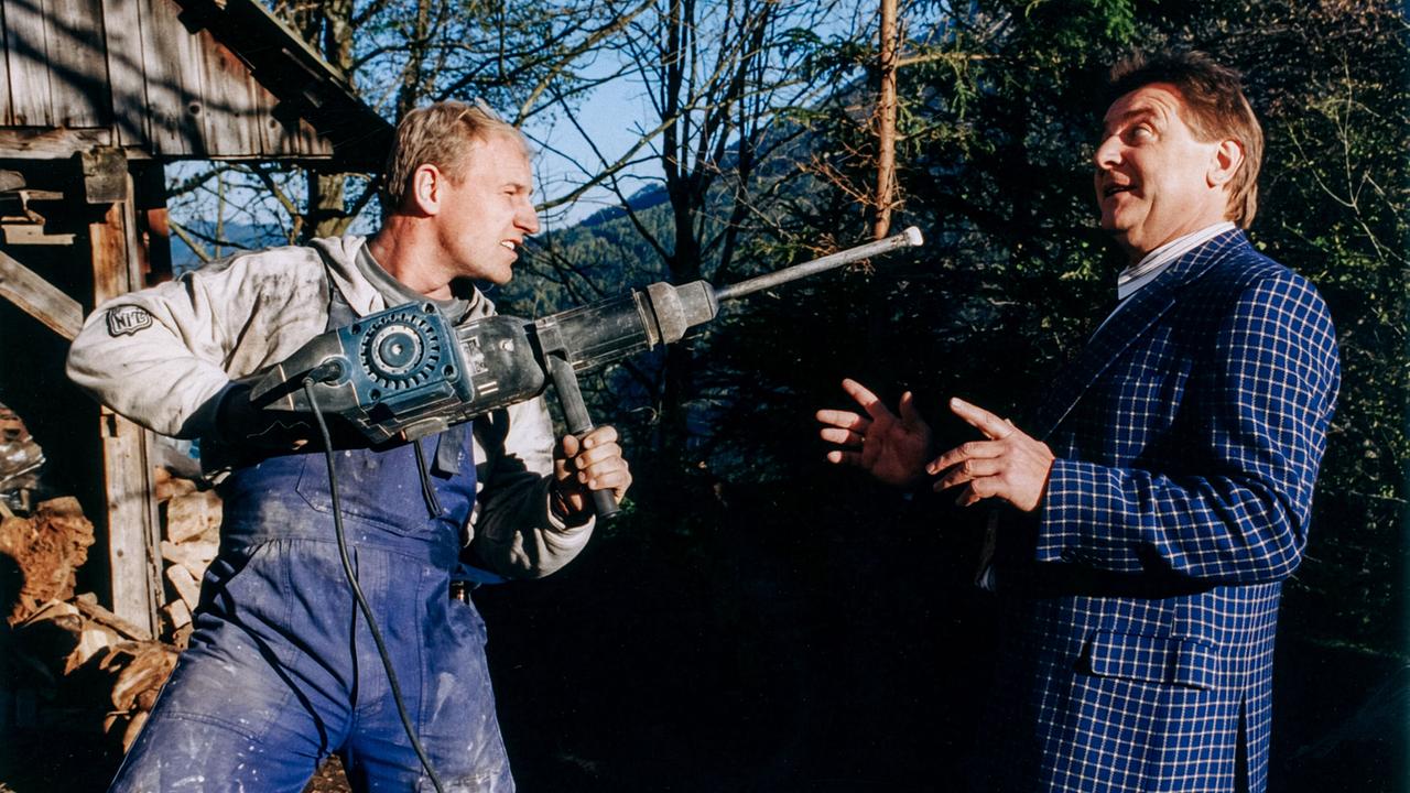 © ORF/DOR Film/Petro Domenigg Herbert Kracal im Handwerker-Outfit bedroht Willi im karierten Sakko im Freien mit einer großen Bohrmaschine.