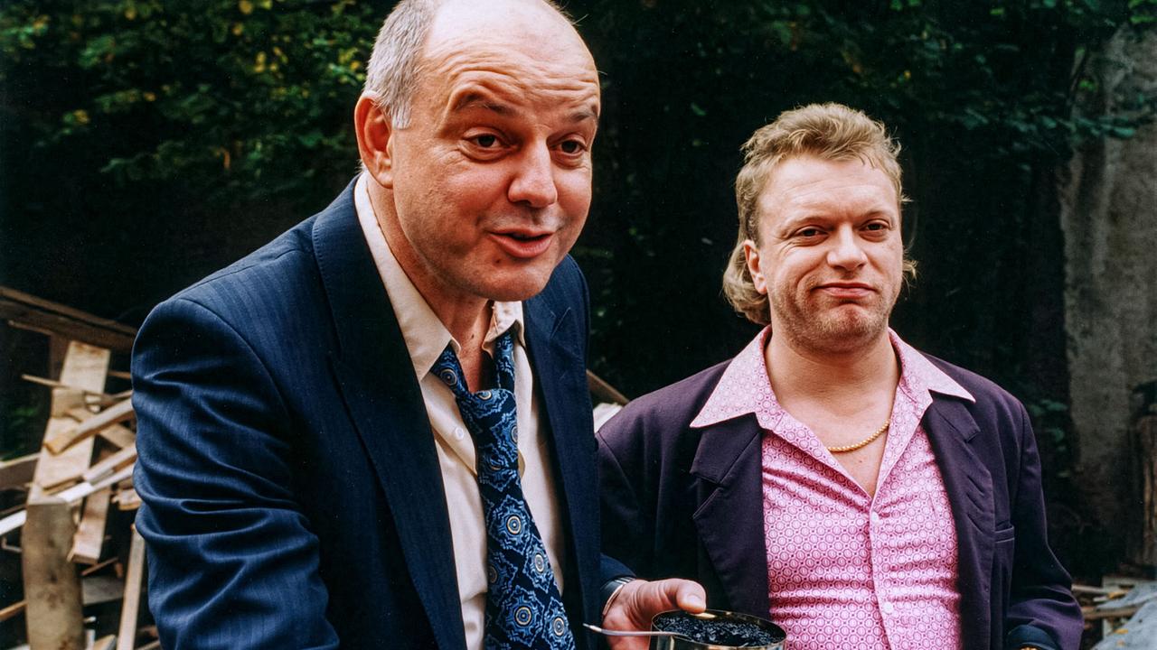 © ORF/DOR Film/Petro Domenigg Meier mit einer Dose Kaviar in der Hand und Sepp stehen im Freien vor Bauholz und dichtem Gebüsch.