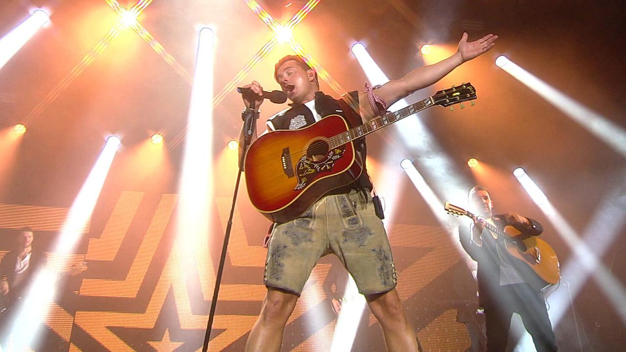 © ORF/Constanze Grießler Andreas Gabalier in Lederhose, weißem T-Shirt und ärmelloser Jacke, mit umgehängter Gitarre und Mikrofonständer in der Hand singt er auf der hell beleuchteten Bühne
