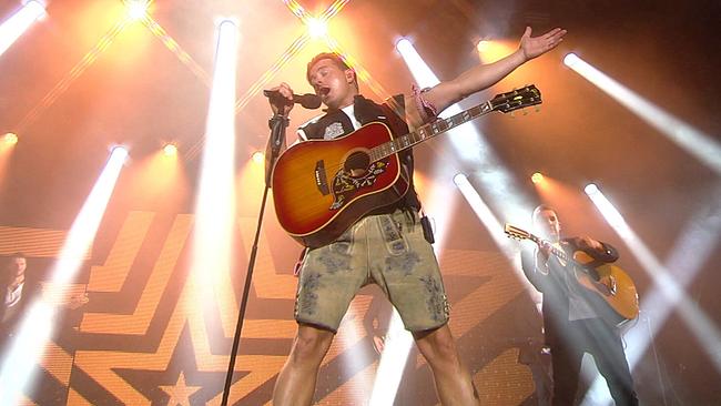 © ORF/Constanze Grießler Andreas Gabalier in Lederhose, weißem T-Shirt und ärmelloser Jacke, mit umgehängter Gitarre und Mikrofonständer in der Hand singt er auf der hell beleuchteten Bühne