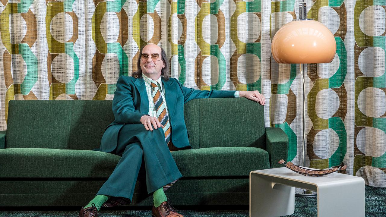 © ORF/dsa gmbh/m. claushallmann Guildo Horn in grünem Anzug, kariertem Hemd, gestreifter Krawatte sitzt er mit Brille auf einem grünen Sofa vor großgemustertem Vorhang im 1970er Ambiente
