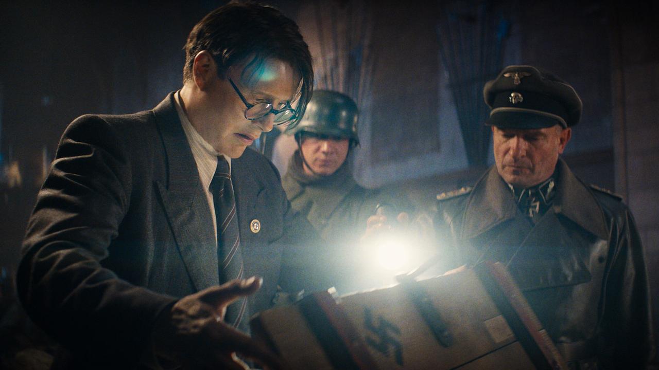 Jürgen Völler mit zwei Männern in NS-Uniformen, in einem dunklen Raum leuchten sie mit einer Taschenlampe in eine Kiste mit Hakenkreuzsymbol, um deren Inhalt zu untersuchen