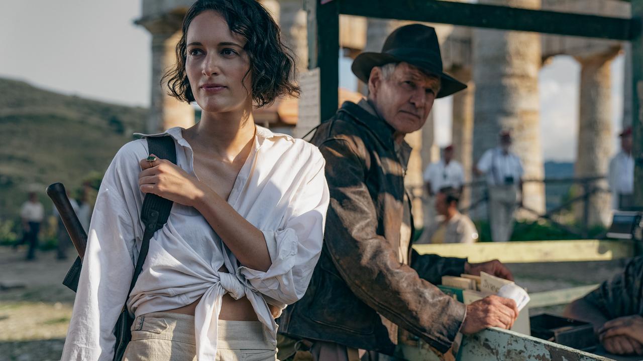 Eine Frau mit weißem Hemd und Rucksack steht im Vordergrund, hinter ihr Indiana Jones mit braunem Hut und Lederjacke - vor sonniger Kulisse eines antiken Tempels