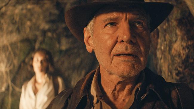 Harrison Ford als Indiana Jones mit braunem Hut und Lederjacke im Vordergrund, dahinter unscharf eine Person in heller Kleidung vor einer Felswand
