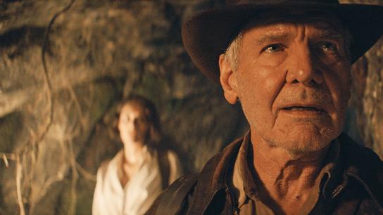 Harrison Ford als Indiana Jones mit braunem Hut und Lederjacke im Vordergrund, dahinter unscharf eine Person in heller Kleidung vor einer Felswand