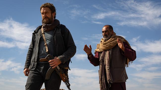 Zwei Männer stehen unter blauem Himmel in einer Wüstenumgebung, einer trägt ein Gewehr, beide sind in Jacken, Schals und robuste Kleidung gehüllt