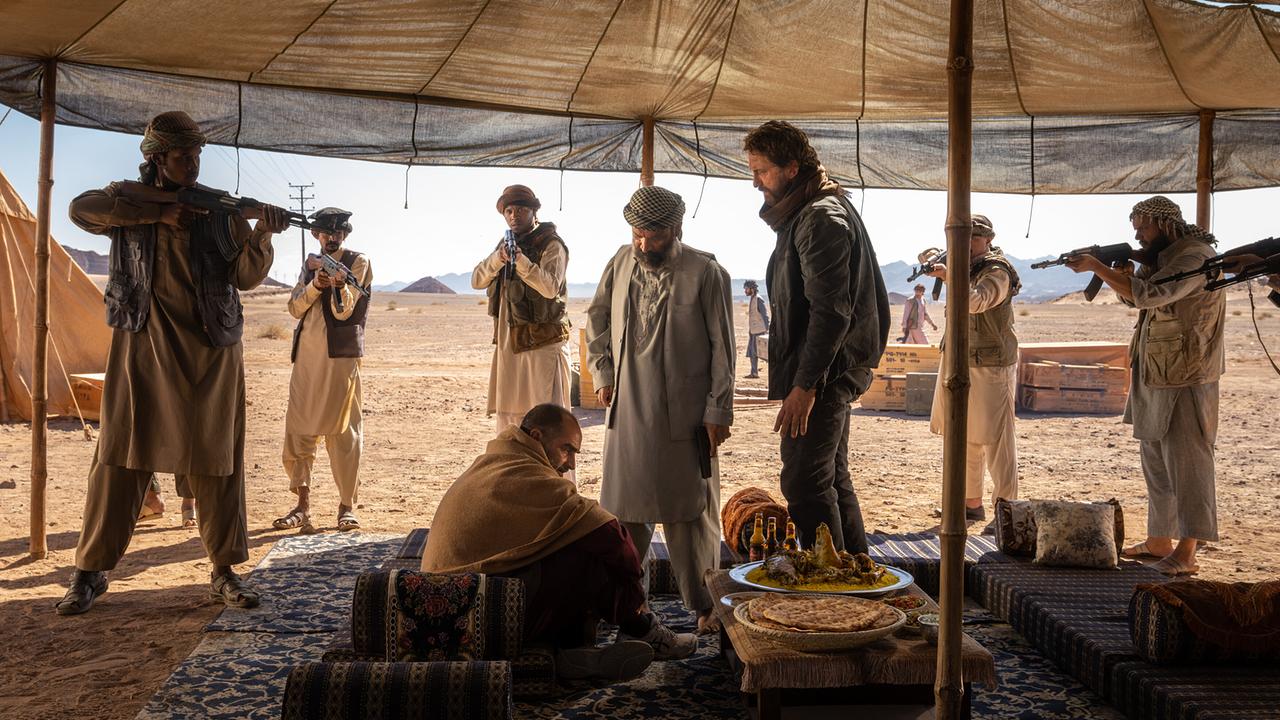 Unter einem Zeltdach in einer Wüstenlandschaft stehen mehrere bewaffnete Männer, während drei Männer im Vordergrund um einen niedrigen Tisch sitzen oder stehen