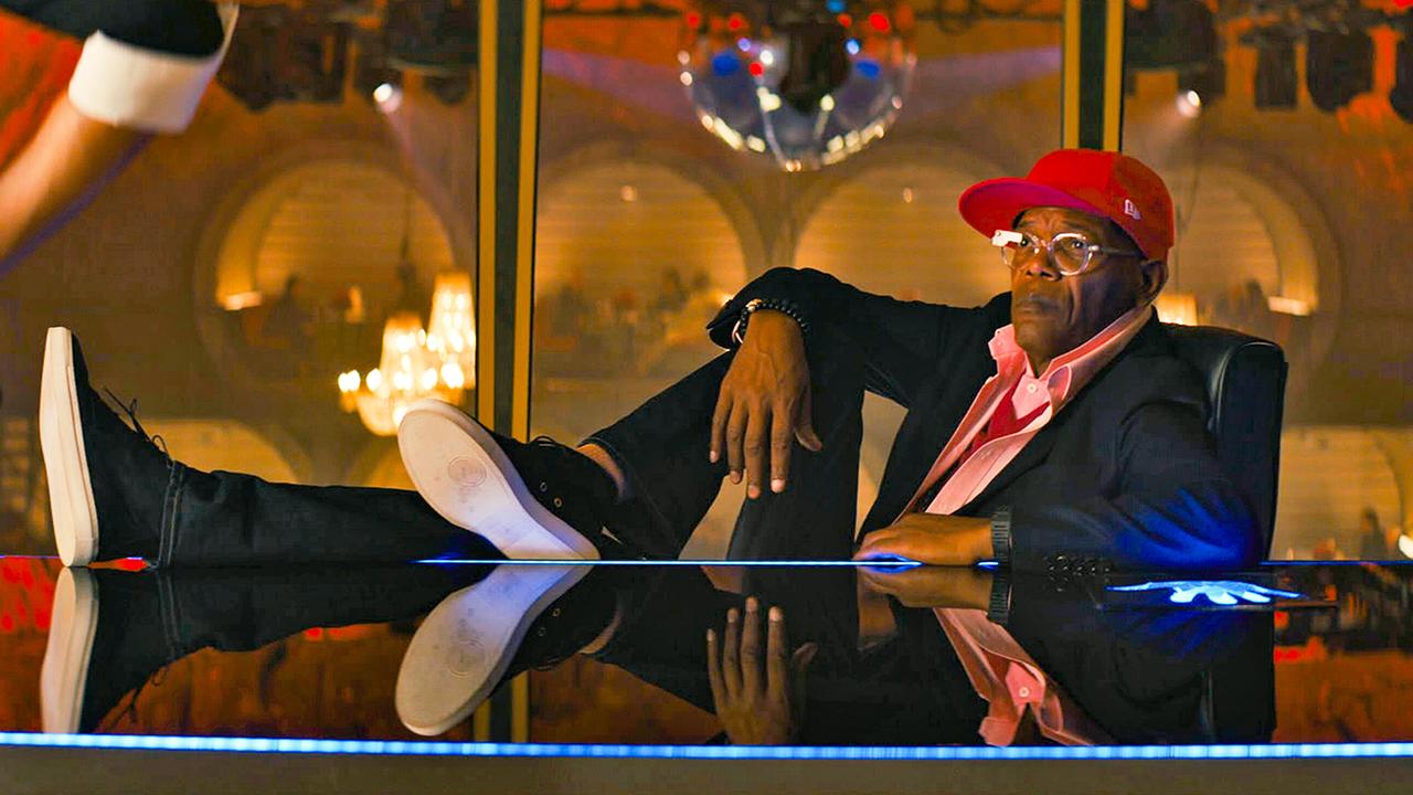 Samuel L. Jackson als Valentine im schwarzen Anzug, mit lachsfarbenem Hemd und roter Schirmmütze, mit seinen Sneakers auf dem modernen Schreibtisch lehnt er lässig in seinem Chefsessel, im Hintergrund ein Raum mit Discokugel